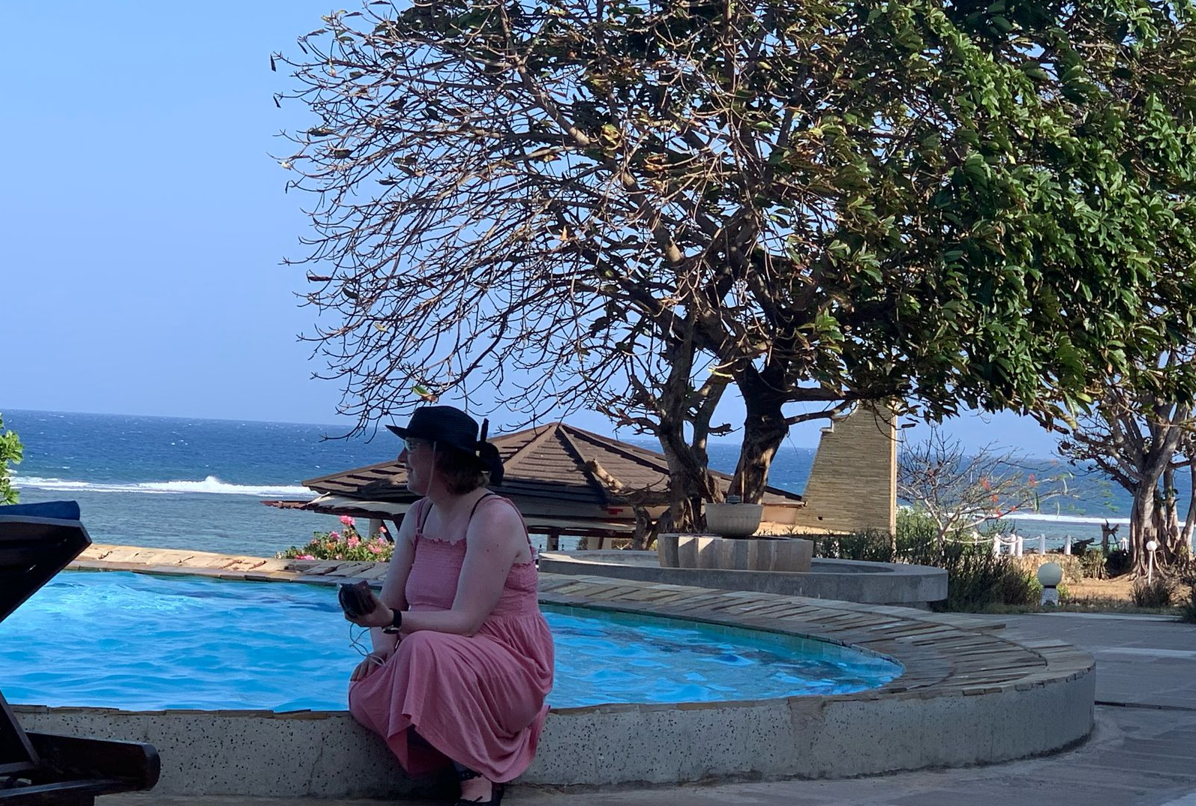 Foto van Lonneke aan de rand van een zwembad met uitzicht op zee. Aan de andere kant van het zwembad staat een grote boom. Lonneke draagt een zomerse, lichtroze jurk en een zwarte hoed. 
